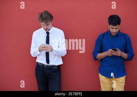 Middle Eastern and caucasian men using smartphones leaning against red wall outdoors Stock Photo