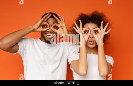 Cheerful Black couple showing okay symbol around eyes while standing against orange background in studio Stock Photo