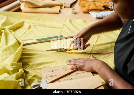 Cropped image of Black female fashion designer sitting cutting yellow fabric using scissors on table with various fabrics, ruler, pencil, and fashi... Stock Photo