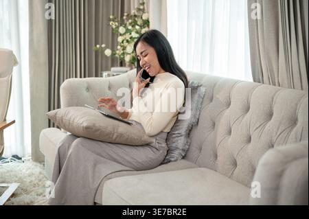 Asian woman having phone conversation while using tablet sitting on sofa in living room at home Stock Photo