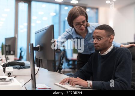 Serious Black man sitting at desk working on his computer talking to caucasian female co-worker leaning over his shoulder pointing something on the... Stock Photo