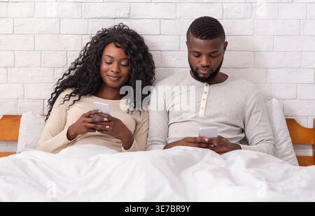Black couple using smartphones sitting together under white blanket on bed in bedroom at home Stock Photo