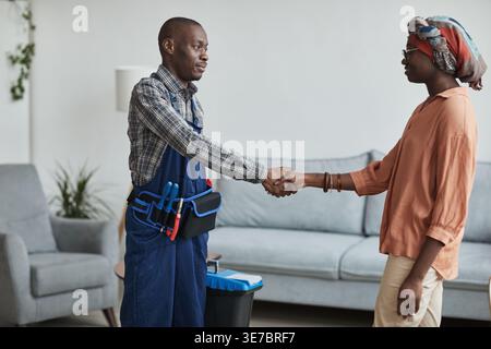 Black handyman wearing tool belt holding toolbox shaking hands with female client while standing at home Stock Photo