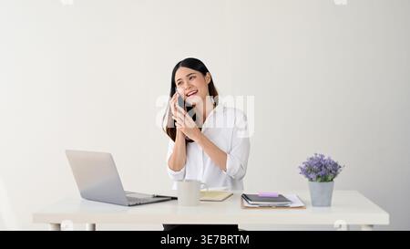 Smiling Asian woman having phone call while sitting at table with laptop against white background in studio Stock Photo