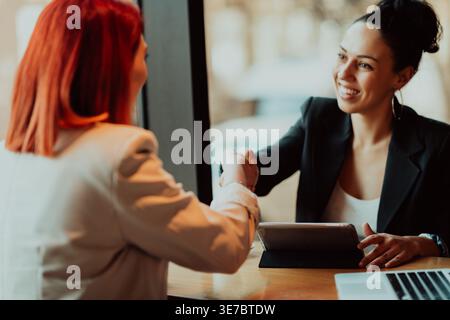 Smiling Hispanic business woman shaking hands with her female colleague while sitting using tablet at table in cafe Stock Photo