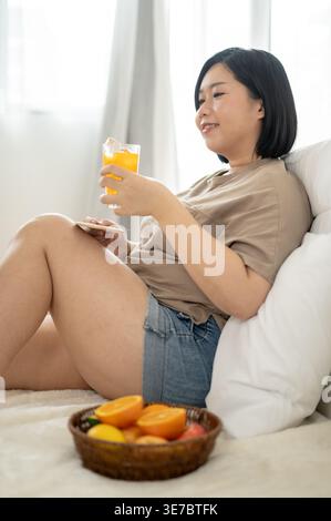 Smiling plus sized Chinese woman holding glass of orange juice and biscuit while sitting on sofa at home Stock Photo