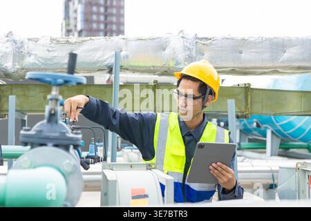 Asian male worker wearing safety helmet checking pipeline using tablet crouching on rooftop during daytime Stock Photo