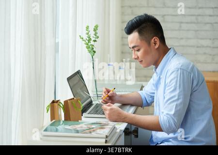 Asian businessman holding pen and paper checking orders made on his online handmade soap shop while sitting at table with laptop at home Stock Photo