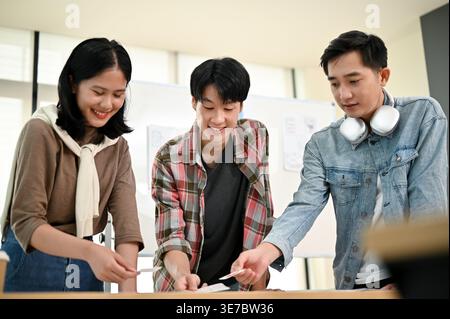 Smiling Asian male and female graphic designers standing at table brainstorming and planing a new project together during team meeting in office Stock Photo