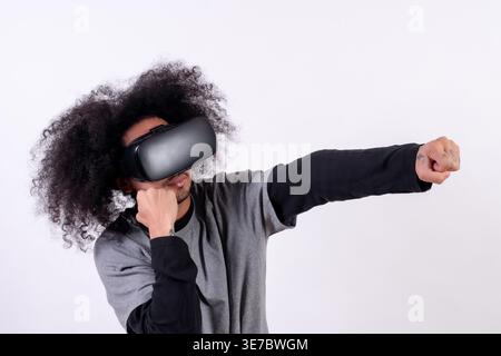 Hispanic man with curly hair standing while wearing VR glasses with clenched fists against white background in studio Stock Photo