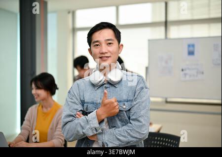 Smiling Asian male office worker wearing headphones around neck showing thumbs up while standing with crossed arms near colleagues working in office Stock Photo