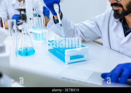 Indian male scientist working with chemical liquids in flask near caucasian female colleague using microscope in laboratory Stock Photo