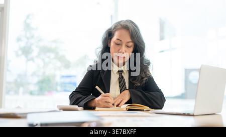 Serious senior Asian business woman writing notes while sitting at table working in the office Stock Photo