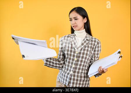 Stressed Asian business woman holding and looking at documents while standing against yellow background in studio Stock Photo