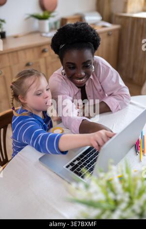Black female tutor pointing on laptop with caucasian girl with down syndrome sitting at table at home Stock Photo
