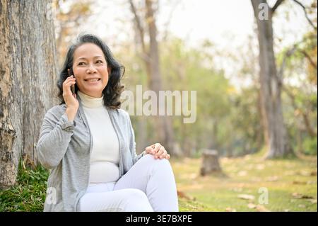 Smiling senior Asian woman looking away having phone conversation sitting on lawn near tree in park during daytime Stock Photo