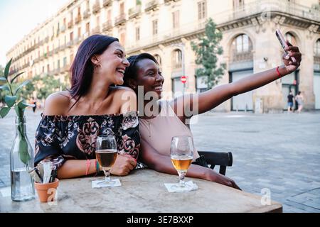 Black woman taking selfie with caucasian female friend sitting at table in outdoor bar in city during daytime Stock Photo