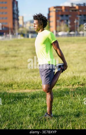 Serious Black man with afro wearing neon green shirt stretching legs looking away while standing in city park on sunny day Stock Photo