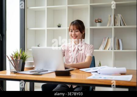 Smiling Asian woman working on laptop sutting at table in office Stock Photo