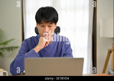 Asian male leaning face on hand  working on laptop at home office during daytime Stock Photo