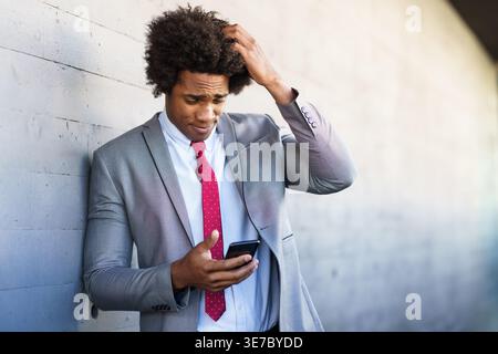 Black businessman with afro hair wearing gray suit and red necktie scratching head using smartphone while leaning against concrete wall outdoors Stock Photo