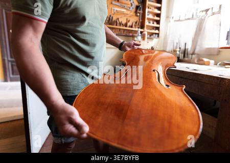 Senior Mixed race male luthier holding polished violin top plate standing near window in workshop Stock Photo