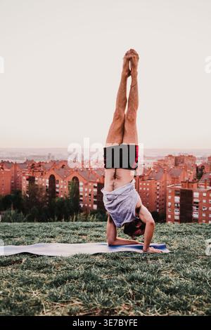 Hispanic man doing handstand on grass outdoors against skyline of residential buildings at sunset Stock Photo