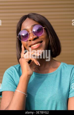 Smiling Black female in sunglasses touching face while standing against brown wall Stock Photo