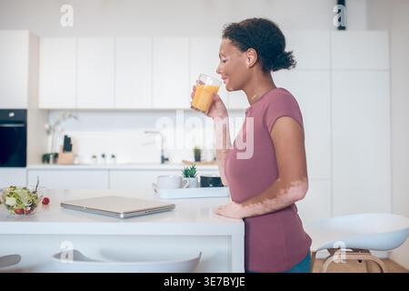 Mixed race woman with vitiligo drinking juice leaning on kitchen counter at home Stock Photo