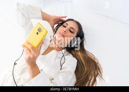 Serious Hispanic woman wearing bathrobe and headphones connected to smartphone listening to music while lying on bed in the hotel Stock Photo
