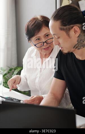 Senior mixed-race mother asking help from his son using laptop while sitting at table with calculator in living room at home Stock Photo