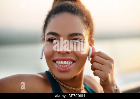 Smiling Mixed race woman touching wireless earphones looking away standing and leaning on railing on promenade during daytime Stock Photo