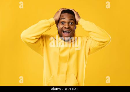 Shocked bearded Black man wearing hoodie touching head with both hands while standing with mouth opened in front of yellow background in studio Stock Photo