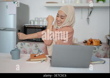 Smiling Asian woman in hijab stretching arms while sitting at table with laptop and books at home Stock Photo