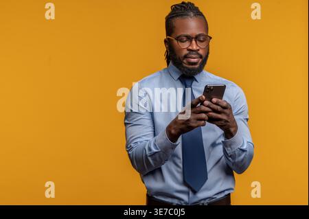 Serious Black man with eyeglasses using smartphone standing against yellow background Stock Photo