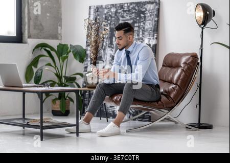Serious Arab businessman using smartphone sitting on leather chair near table with laptop in office Stock Photo