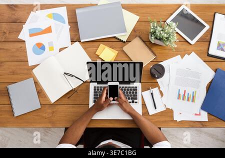 Black businessmen using smartphone sitting at wooden desk with laptop and various objects in office Stock Photo