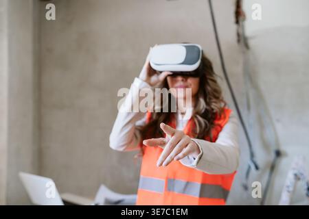 Hispanic female architect using VR headset pointing forward standing in construction site during daytime Stock Photo