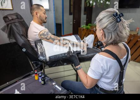 Asian male client sitting on chair looking away and getting tattooed on his arm by middle-aged caucasian female tattoo artist in studio Stock Photo