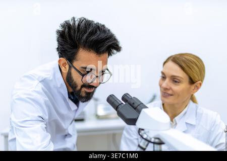 Smiling Indian male and caucasian female scientists wearing lab coats and latex gloves using microscope doing sample analysis together in laboratory Stock Photo