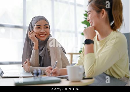 Smiling Asian female friends talking at each other while sitting at table with laptop at home Stock Photo