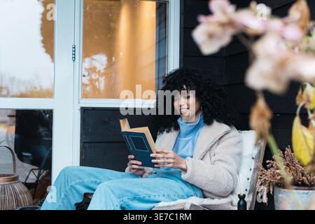 Smiling Mixed race woman with curly hair reading book while sitting on chair on terrace at home Stock Photo