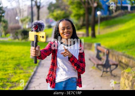 Cheerful Black female social media influencer holding selfie stick recording video using smartphone with mic while walking in park on sunny day Stock Photo