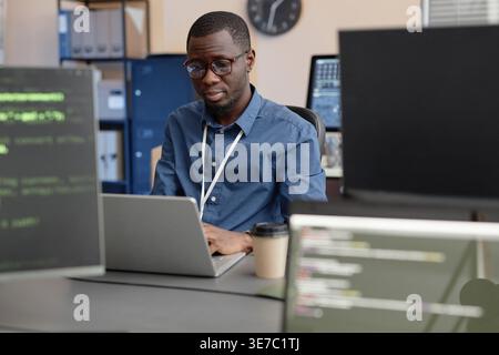 Focused Black male IT specialist wearing eyeglasses using laptop sitting at desk surrounded by computers in office Stock Photo