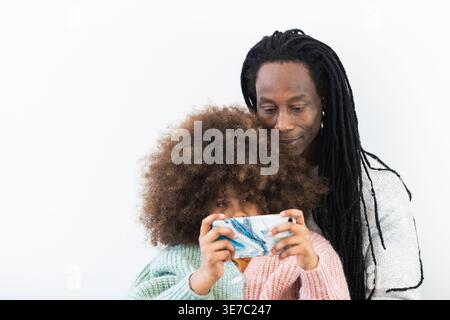 Black father with locks hugging Mixed race daughter with afro while sitting and watching video on smartphone against white background in studio Stock Photo