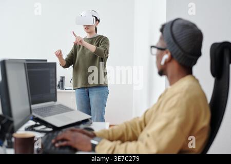 Black man wearing eyeglasses and earphones working on computer looking at Mixed race female colleague with VR headset while sitting at table in office Stock Photo