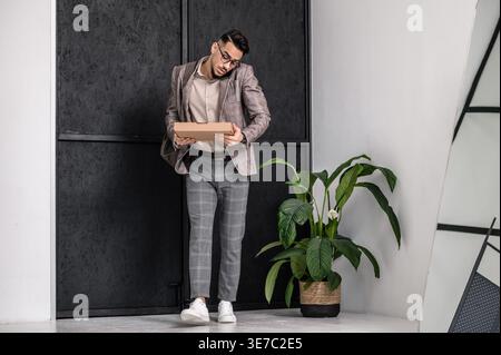 Arab man wearing eyeglasses having phone conversation holding box walking in apartment Stock Photo