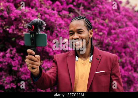 Smiling Hispanic male vlogger with braids recording video using smartphone standing in garden full of flowers during daytime Stock Photo