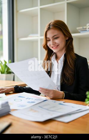 Smiling Asian business woman in black suit looking at reports while sitting at table in office Stock Photo