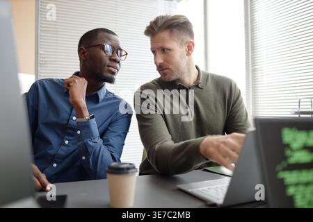 Black male IT specialist with eyeglasses leaning on table discussing project with white male coworker pointing at laptop and looking at him sitting... Stock Photo